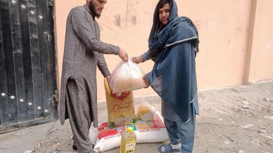 Left: a young man in beige Islamic dress hands a bag of food supplies to a man on the right, wearing blue clothes and head covering.