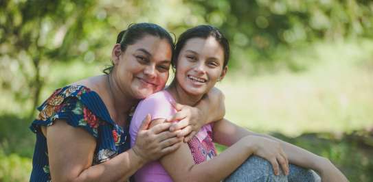 A woman (Angela) sits with her arms round her daughter, who is leaning back into her. They are both smiling at the camera.