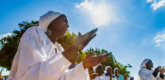 On a sunny day with blue skies, a group of black Zimbabwean women in white religious dress clap and sing