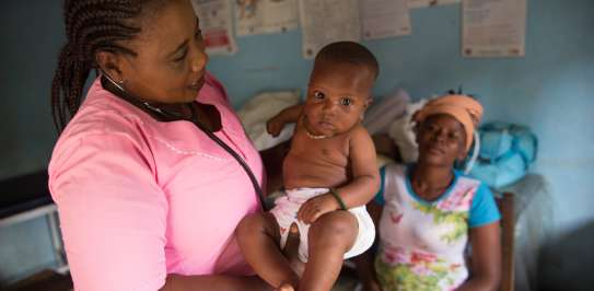 Nurse Judith stands holding a small baby, who is staring at the camera. The baby's mother sits, out of focus, in the background.