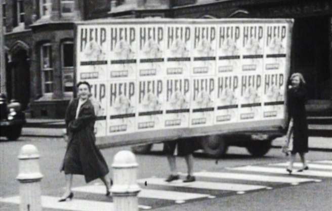 Women cross the road carrying a large banner covered in posters with 'help' written on them.