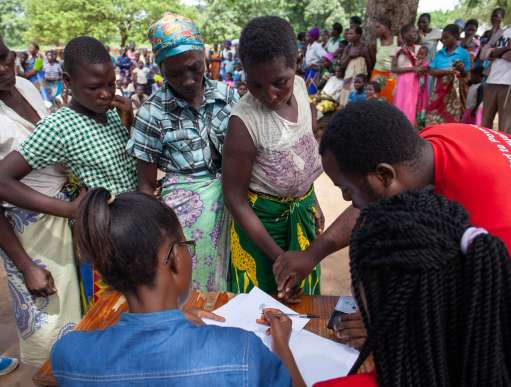 Malawi. A group of black women queue up at a table, behind which 2 black women are seated; a black man in a red t-shirt, to the right of the shot, helps the first woman with her application. 