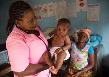 Nurse Judith stands holding a small baby, who is staring at the camera. The baby's mother sits, out of focus, in the background.