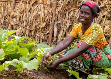 A proudly smiling black woman wearing brightly patterned attire crouches down by her garden of lush green plants with soil in her hands